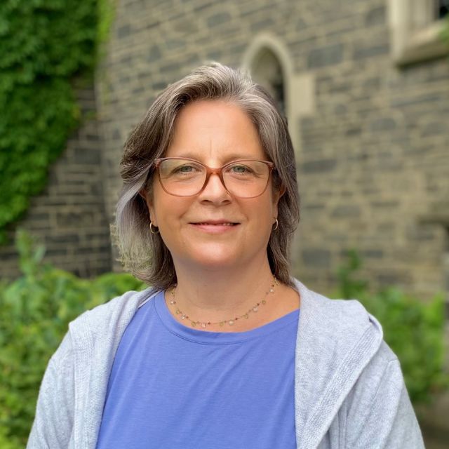 Jennifer is a white woman with greying shoulder length hair. She wears glasses and a blue shirt under a light grey cardigan.